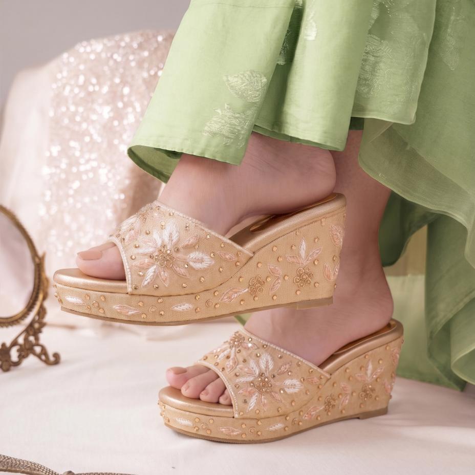 A pair of beige embellished wedge sandals worn with light green embroidered pants, shown on a model's feet against a soft, light-colored background.
