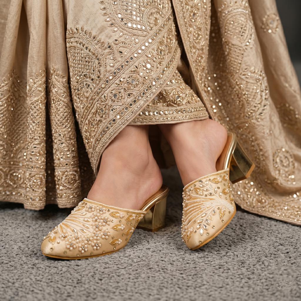 A close-up of a person wearing ornate beige and gold embellished mule heels with a matching embroidered garment draped around the legs, standing on a gray textured floor.