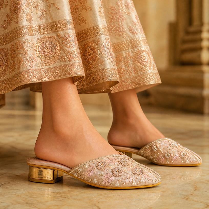 A pair of women's pointed-toe mule heels with ornate floral embroidery and gold-tone block heels, shown worn with a beige embroidered skirt in an indoor setting.