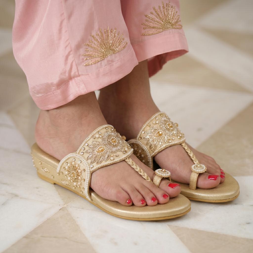 A close-up of a person wearing ornate gold and silver embellished thong sandals with a low wedge heel, paired with light pink embroidered pants, standing on a light marble floor.
