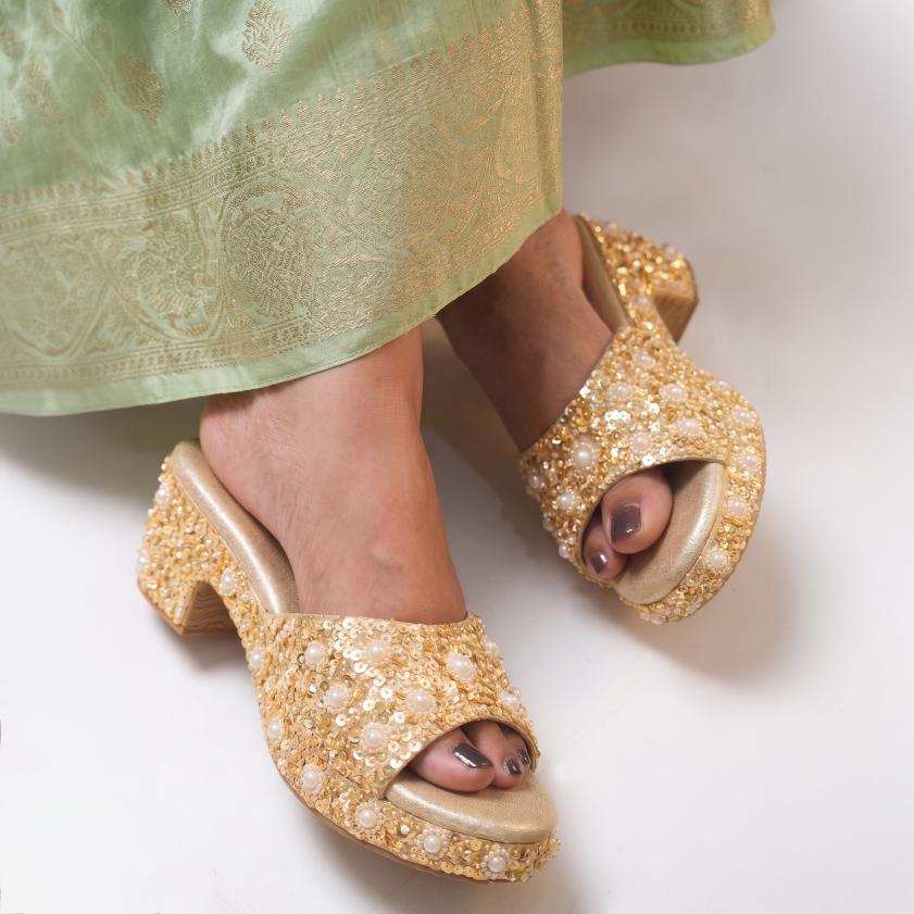 A close-up of a person wearing ornate gold embellished open-toe platform mule sandals with a block heel, paired with a light green patterned skirt.