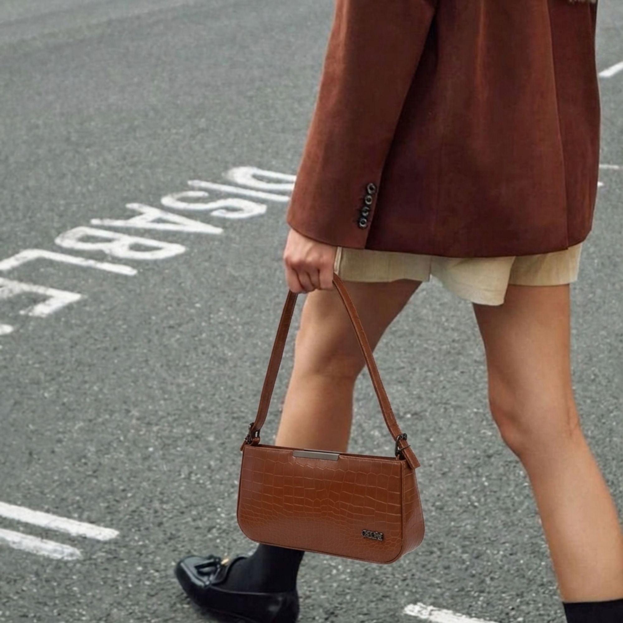 A person walking on an asphalt street carrying a brown crocodile-embossed shoulder bag. The person is wearing a brown suede-like blazer, a cream skirt, and black loafers.
